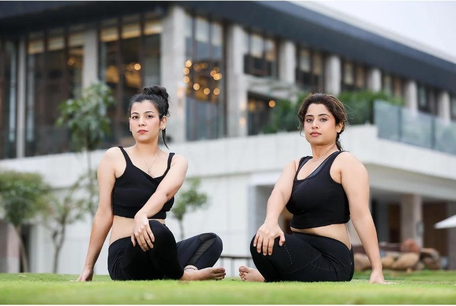 Две жени са седнали на тревна площ и правят стречинг упражнения.